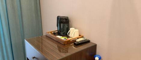 A wooden bedside table with a coffee maker, mugs, a remote control, and a magazine.