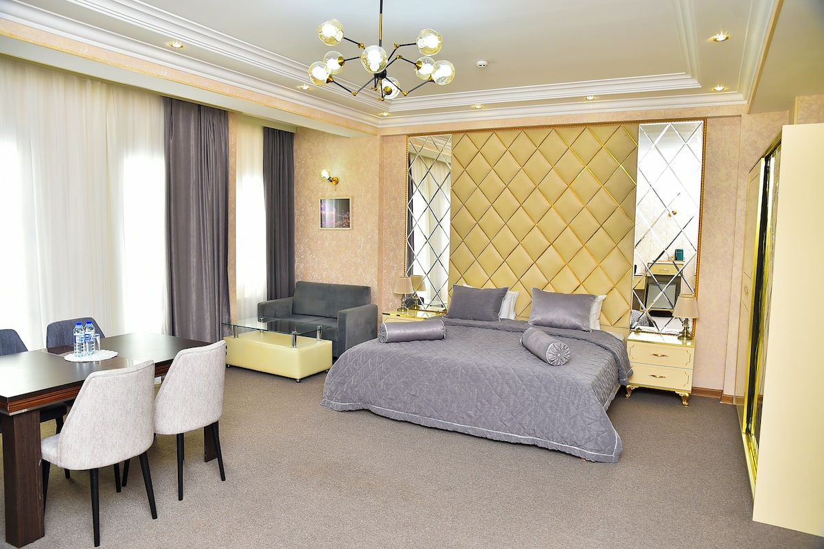 A hotel room with a large bed, a desk with chairs, a sofa, and a bedside table with a lamp.