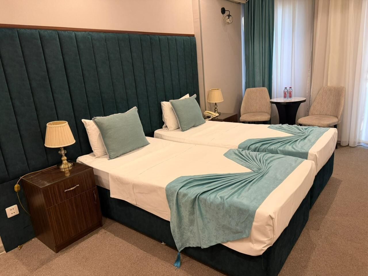 A hotel room with two beds, a nightstand with a lamp, a chair, and a small table with water bottles.