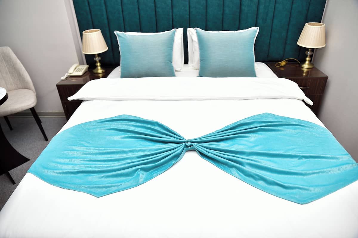 A neatly made bed with a blue bedspread and pillows, a bedside lamp, and a chair.