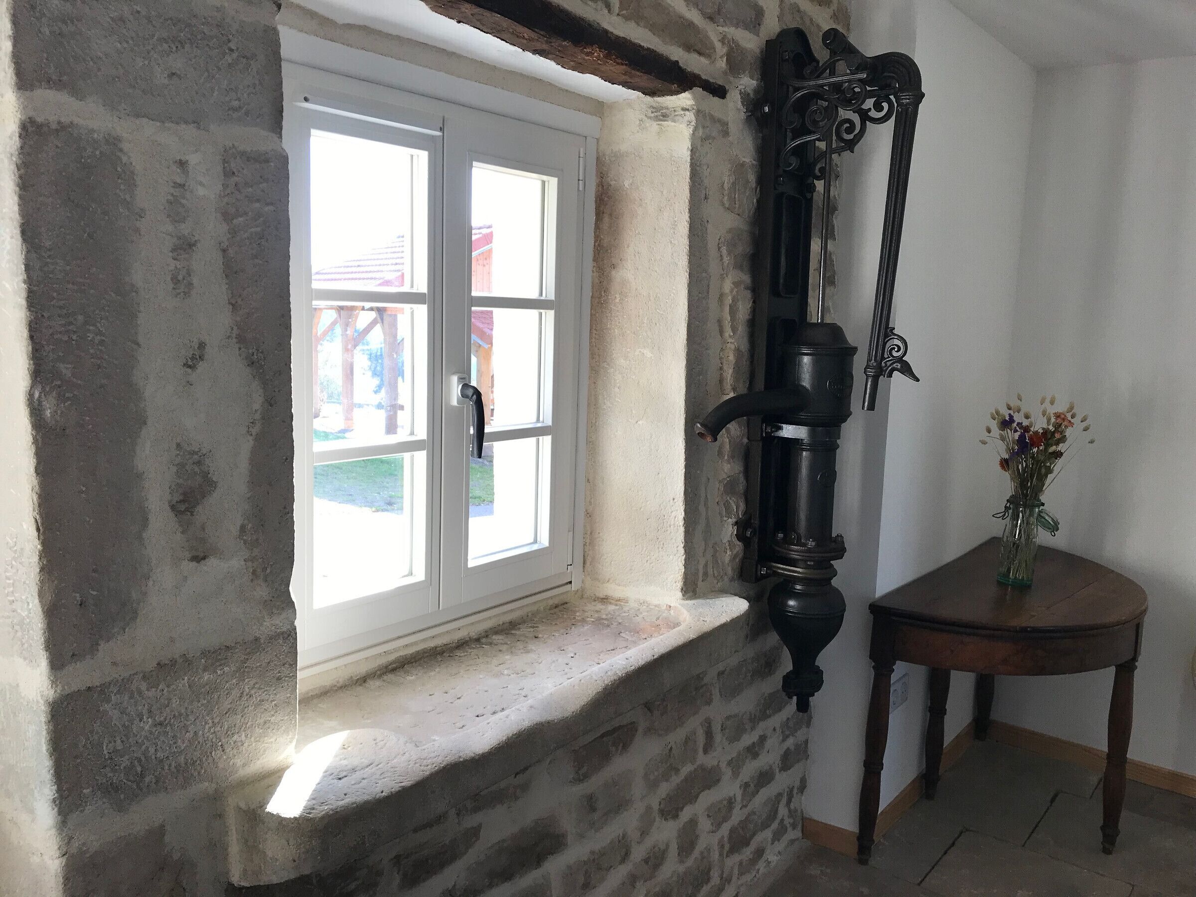 A room with a stone wall, a white-framed window, a wooden table with a vase, and a black wall-mounted object.