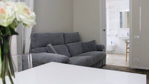 Una habitación moderna con un sofá gris, una mesa blanca y vista a un baño por una puerta abierta.