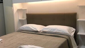 Una cama bien hecha con una colcha color beige y almohadas, una toalla blanca doblada sobre la cama, y un cabecero moderno con iluminación integrada.