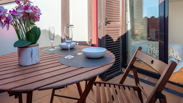 A wooden table with a potted orchid, two wine glasses, and a bowl on a patio with a sliding glass door leading to a bedroom.