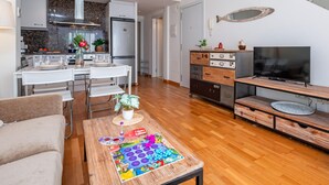 A modern living room with a wooden coffee table, a TV on a wooden stand, a kitchen area with a refrigerator, and a dining table with chairs.