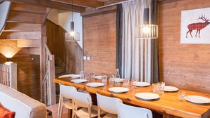A dining area with a long wooden table set for four, white chairs, and a staircase in the background.