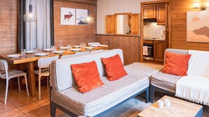 A cozy living area with a dining table set for four, a sofa with orange cushions, and a kitchen area with wooden cabinets and a tiled backsplash.