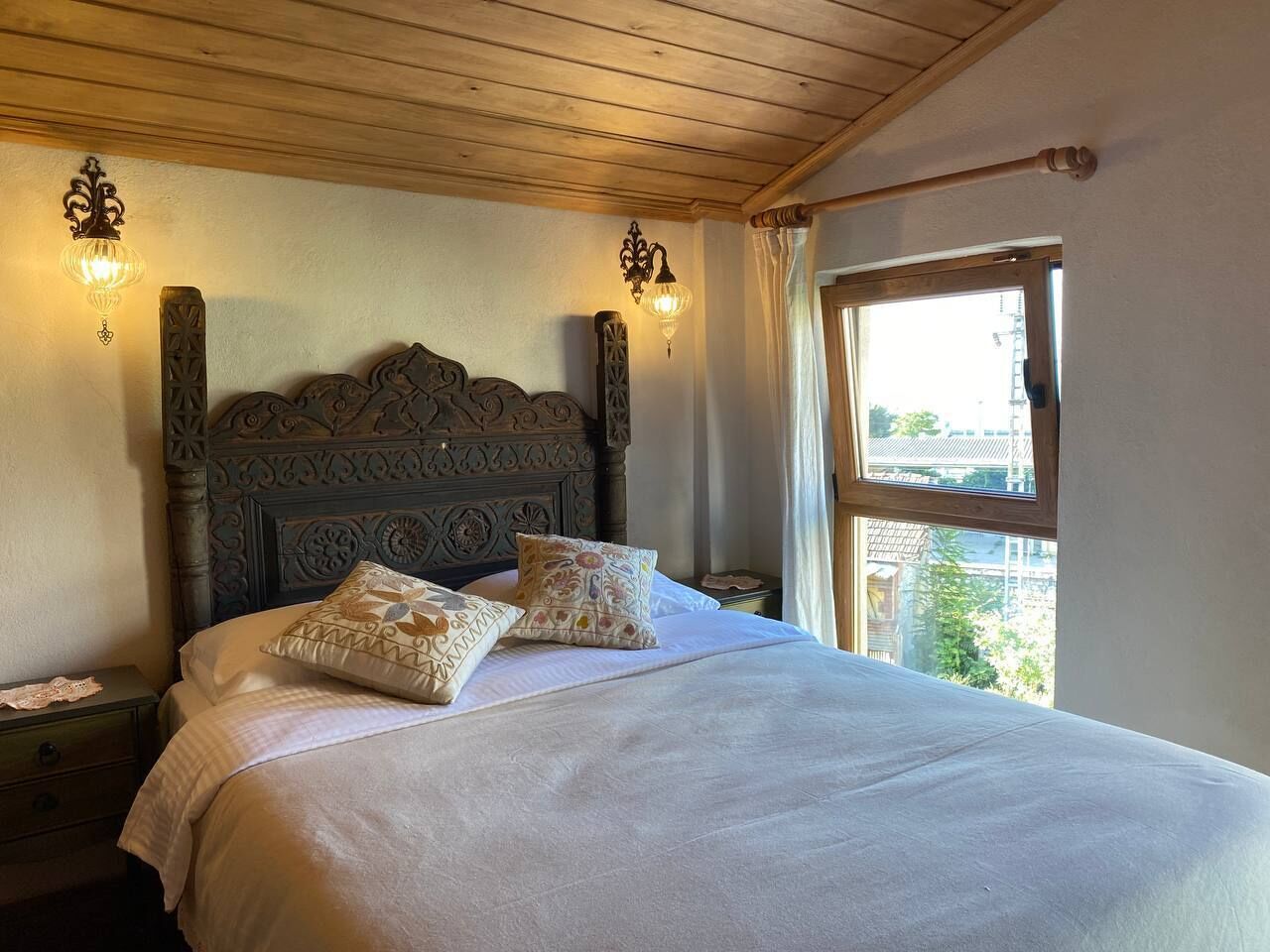 A bed with a wooden headboard, two wall sconces, and a window with curtains.