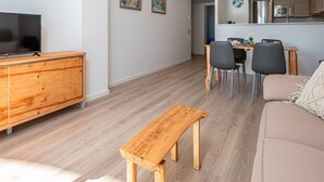 A modern living room with a wooden bench, a sofa, a TV on a wooden cabinet, and a dining area with chairs and a table. - Apartment with Terrace and Parking 5 min from Beach (Palamós)