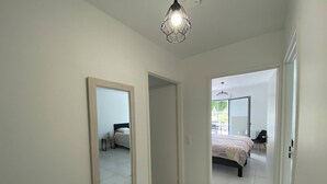 Ein Flur, der zu einem Schlafzimmer führt, mit einem Bett, einem Sessel und einem Tisch. An der linken Wand befindet sich ein Spiegel und auf der rechten eine Tür.