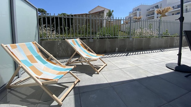 Zwei Holz-Loungestühle mit gestreiften Kissen auf einer gefliesten Terrasse, ein Sonnenschirm und Blick auf eine Wohngegend mit Gebäuden und einem Zaun.