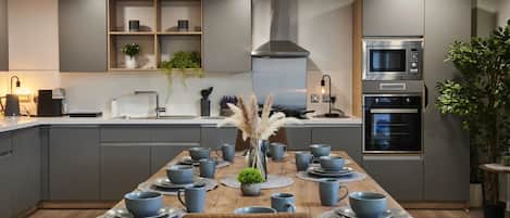 A modern kitchen with grey cabinets, a wooden dining table set with blue cups and saucers, and a central vase with dried plants.