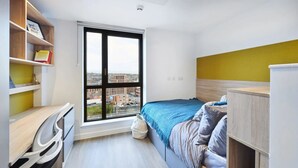 Un dormitori amb una finestra gran, un escriptori, un llit amb llençols blaus i una paret amb tocs de color groc.