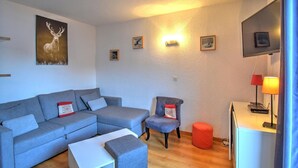 A modern living room with a grey sectional sofa, a small armchair, a white side table, and a red ottoman. There are framed pictures on the wall and a lamp on a side table. - 3-room apartment with southwest view and wifi in Morzine (Morzine)