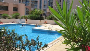 Una piscina amb gent a les proximitats, envoltada d'un edifici amb balcons i vegetació.