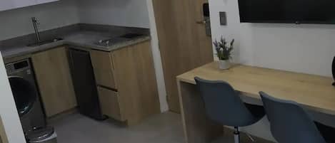 Una habitación de hotel compacta con cama, un escritorio con dos sillas, televisión, una planta pequeña en maceta y una kitchenette con lavamanos y lavadora.