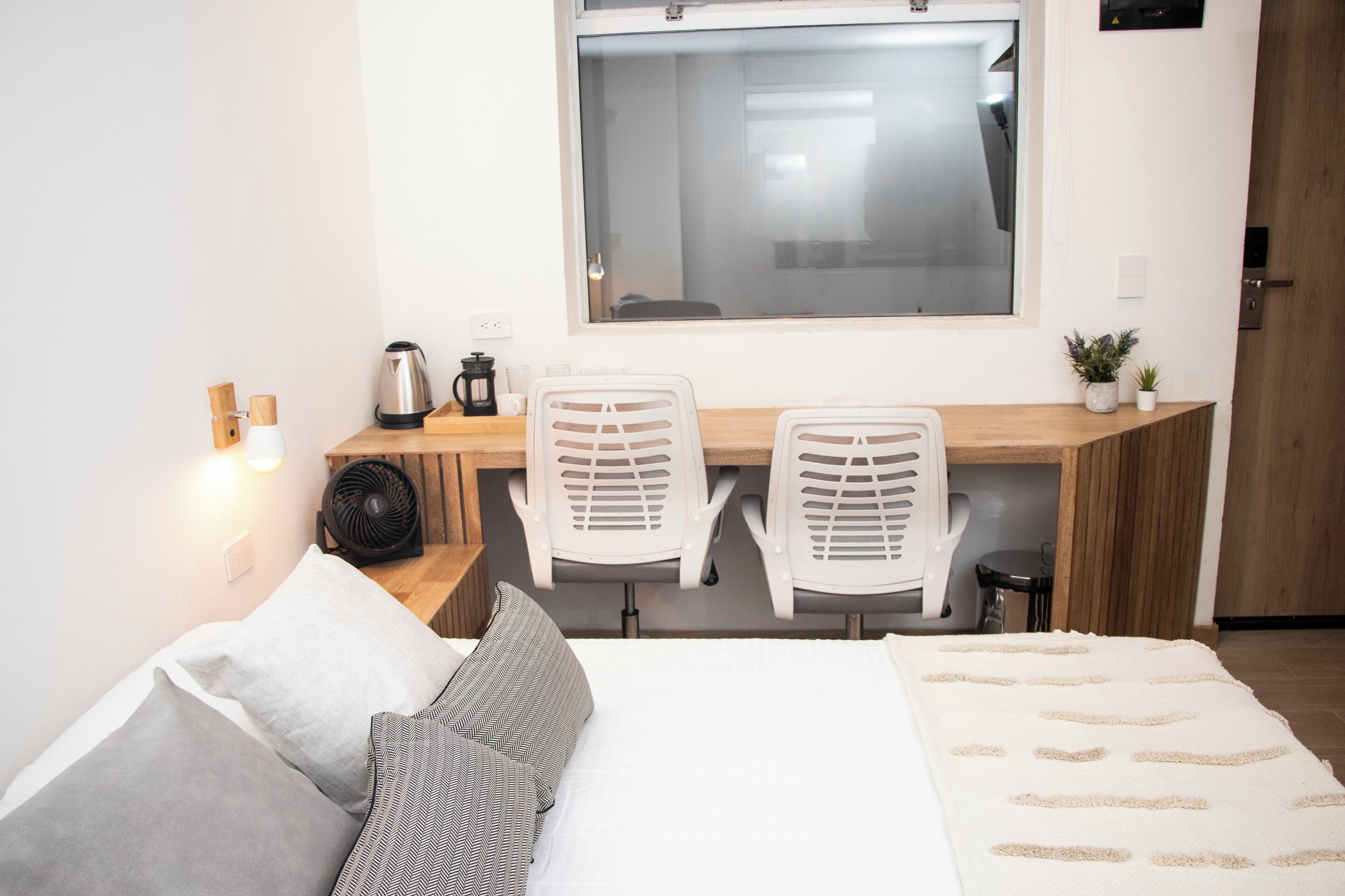 A small, modern room with a bed, two white chairs, a wooden desk, a window, and a small plant.
