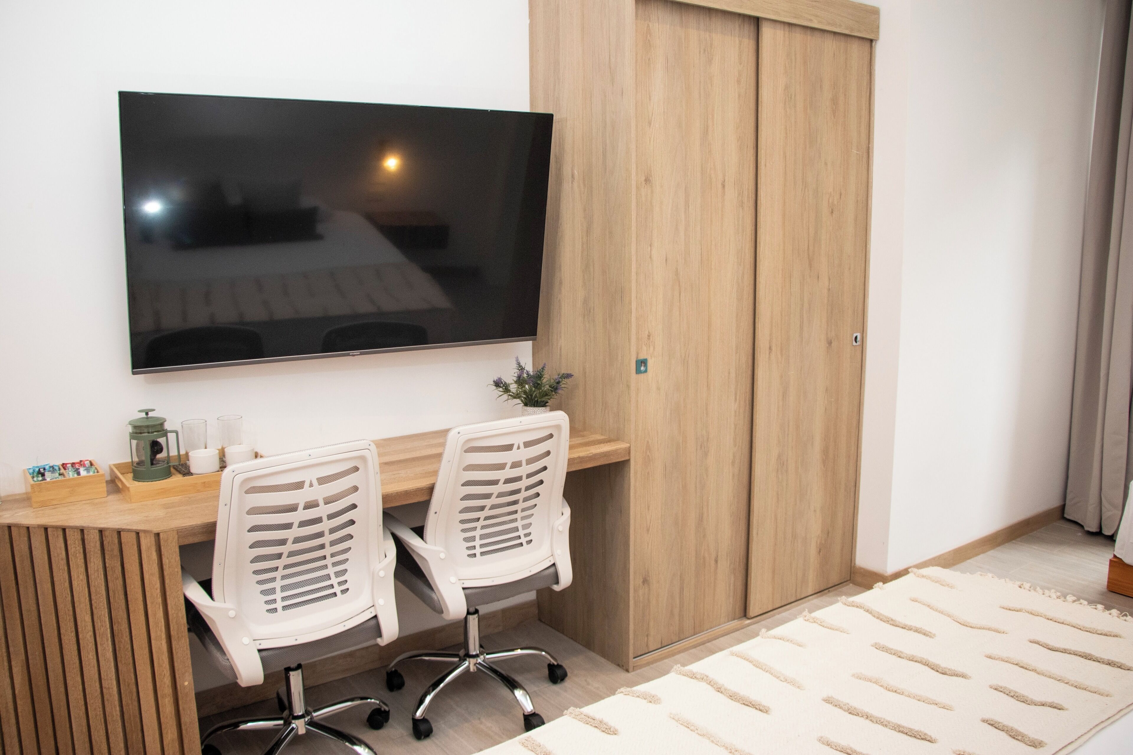 Un espace de bureau moderne avec un grand téléviseur à écran plat fixé au mur, deux chaises de bureau blanches, un bureau en bois et une petite plante.