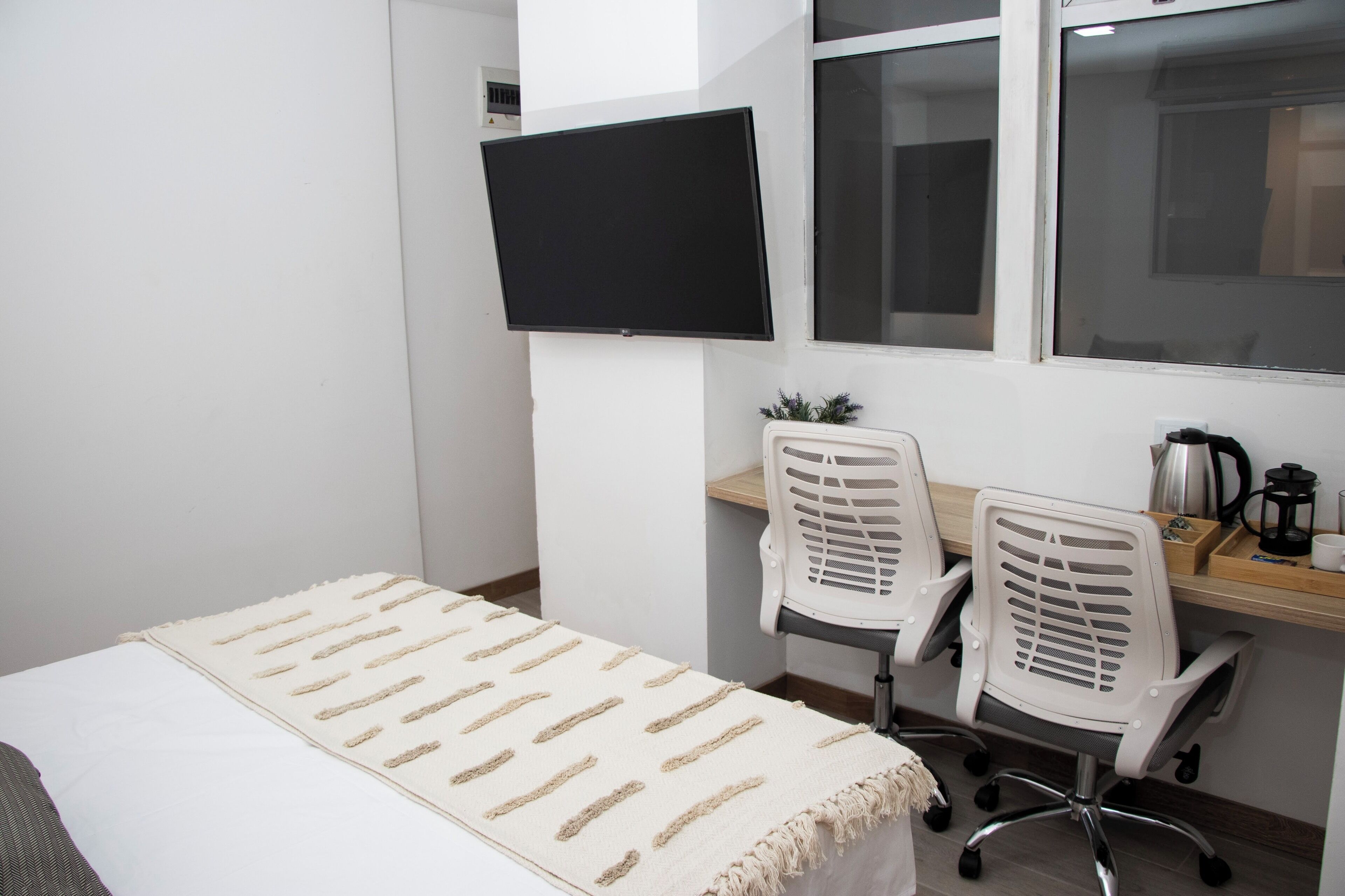 Une petite chambre moderne, équipée d’un lit, d’un bureau avec deux chaises, d’un téléviseur fixé au mur et d’une fenêtre.