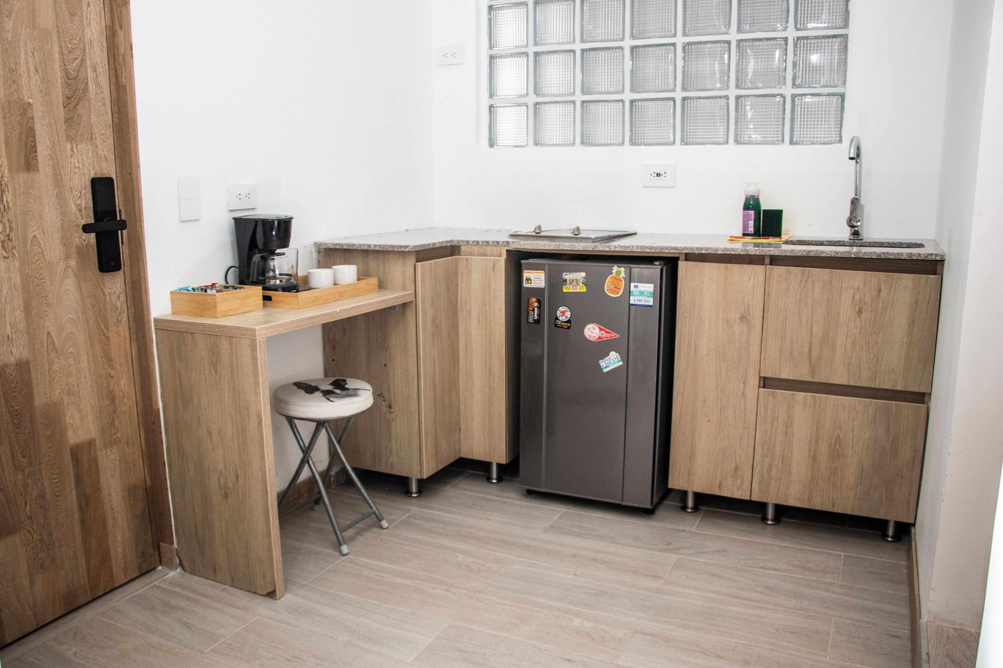 A compact kitchen with wooden cabinets, a stainless steel refrigerator, a coffee maker, and a small stool.