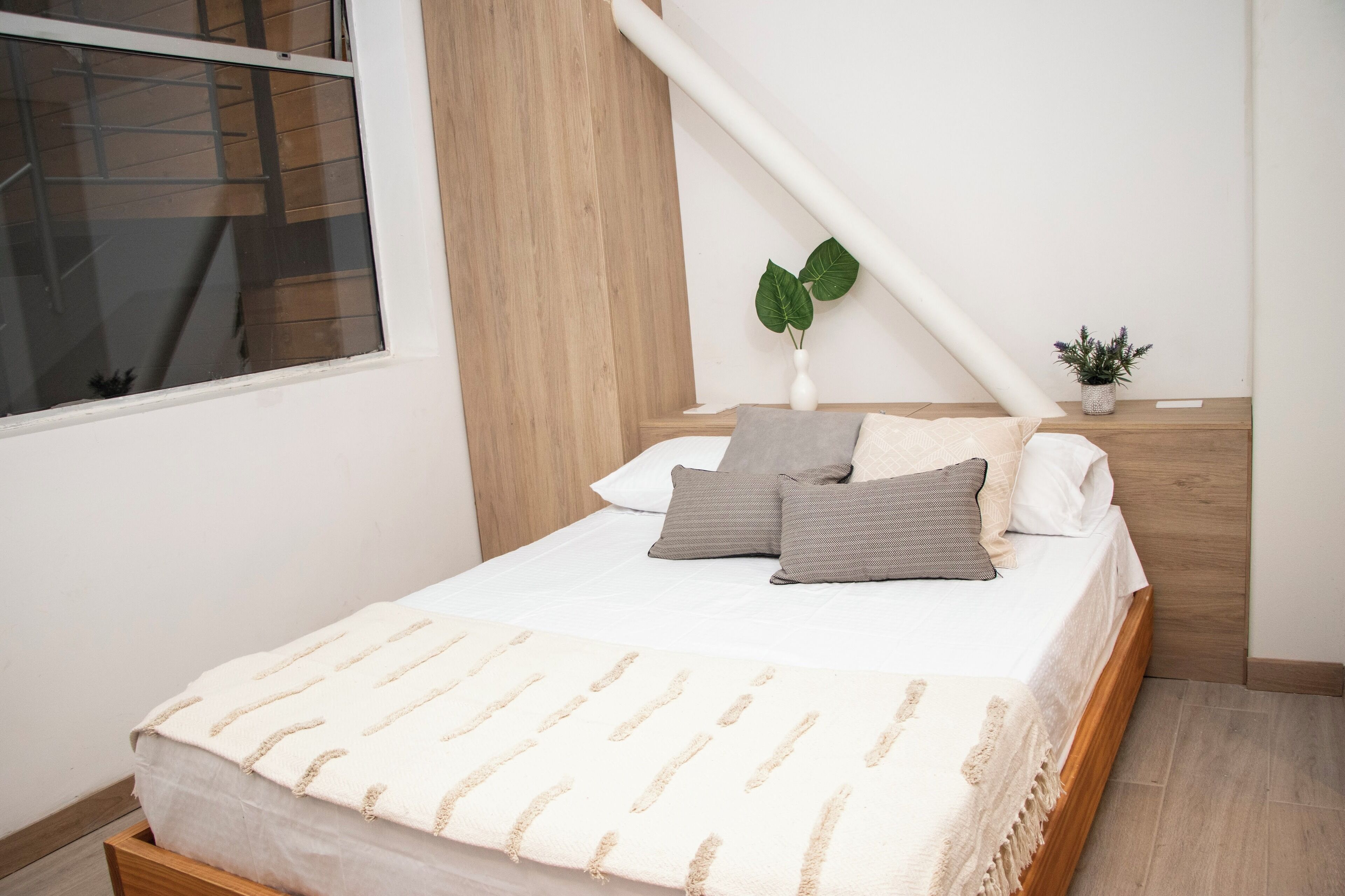 A bedroom with a bed, pillows, a bedside plant, and a window.