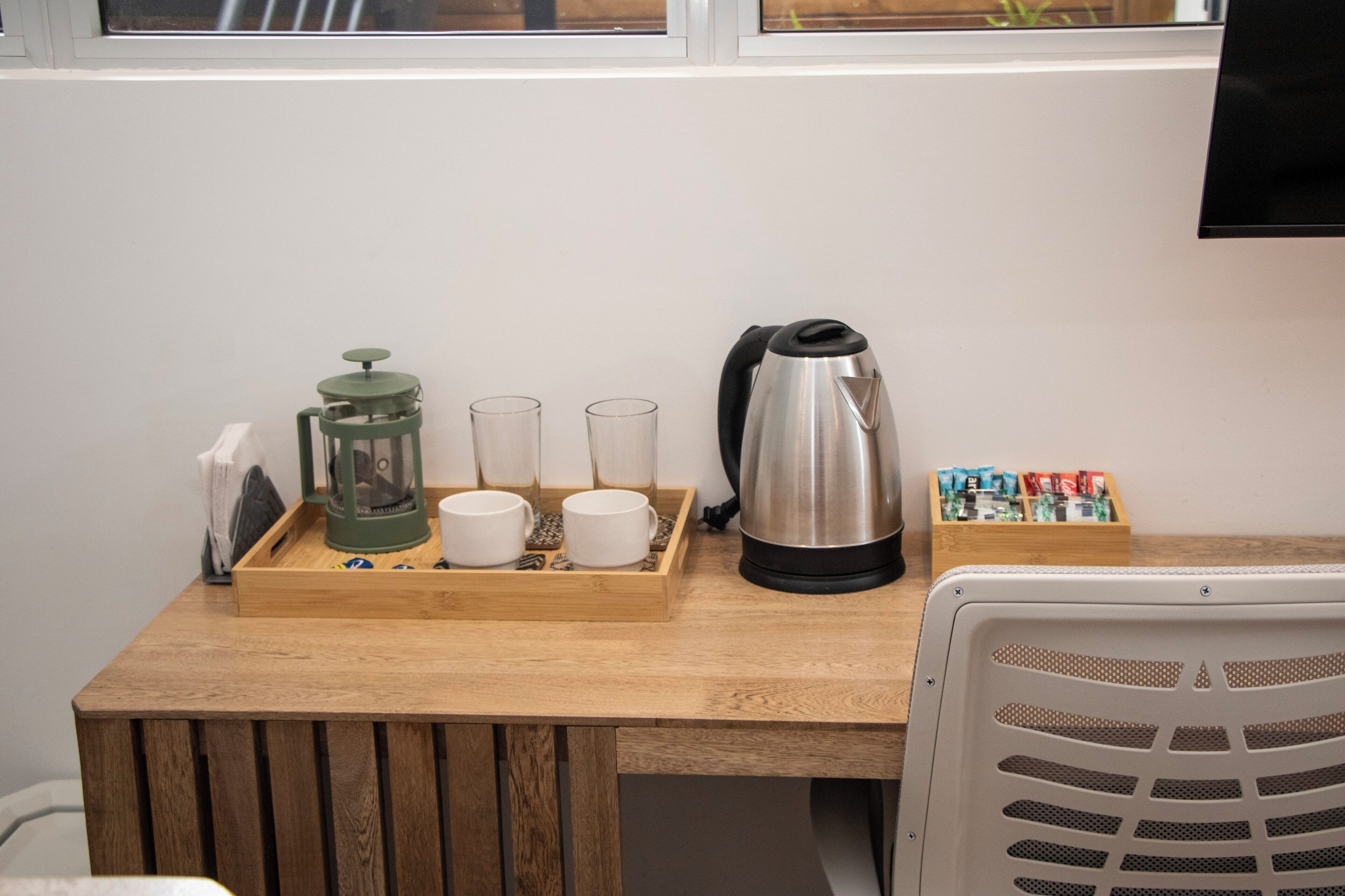 A wooden table with a tray holding a green coffee grinder, two white cups, two glasses, a silver electric kettle, and a box of tea bags.