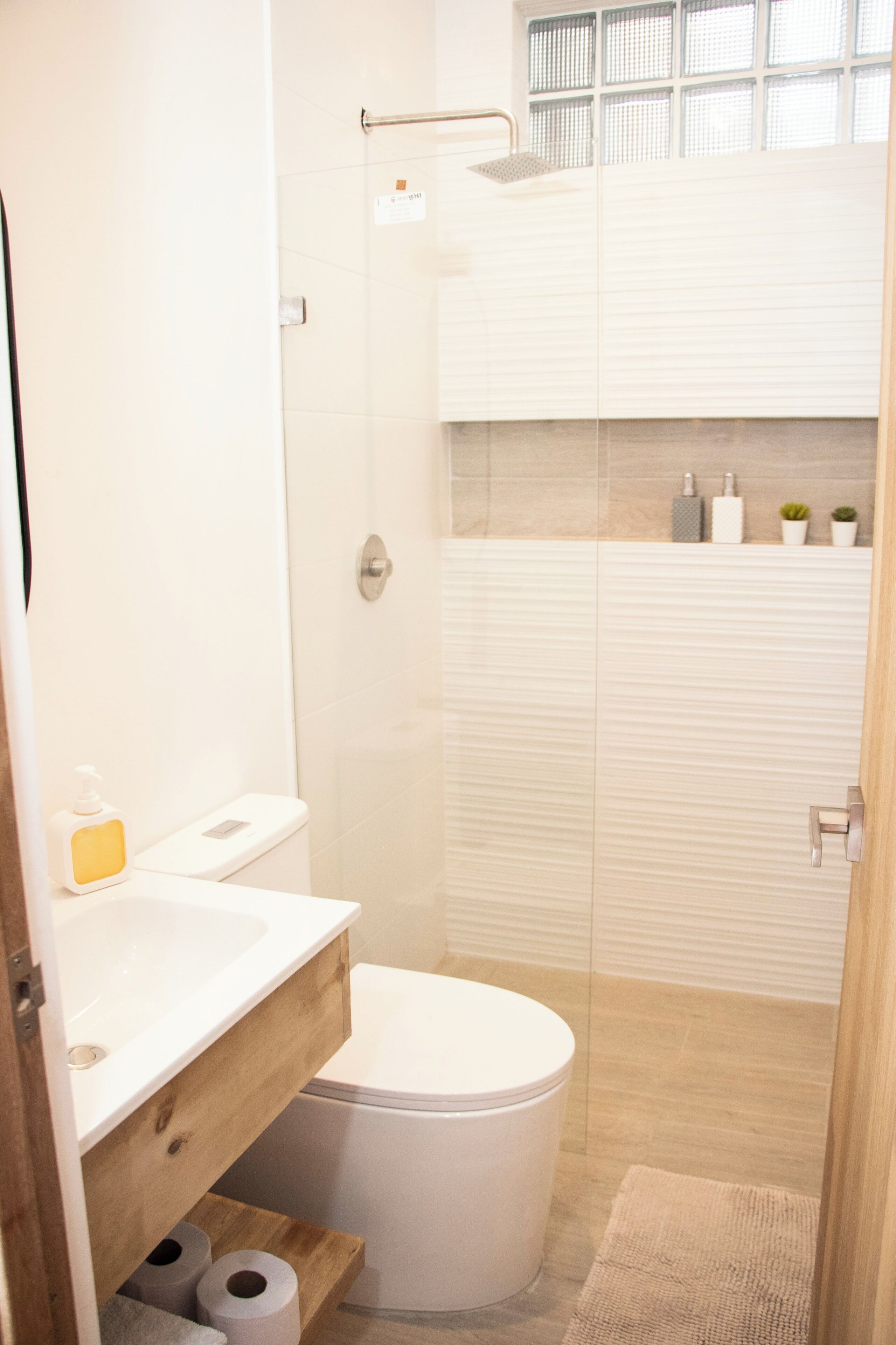 A modern bathroom with a white sink, toilet, and shower. There are toiletries on the sink and a small plant on the wall.