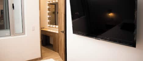 A room with a large flat-screen TV mounted on the wall, a vanity with a mirror and lights, and a door leading to another room.