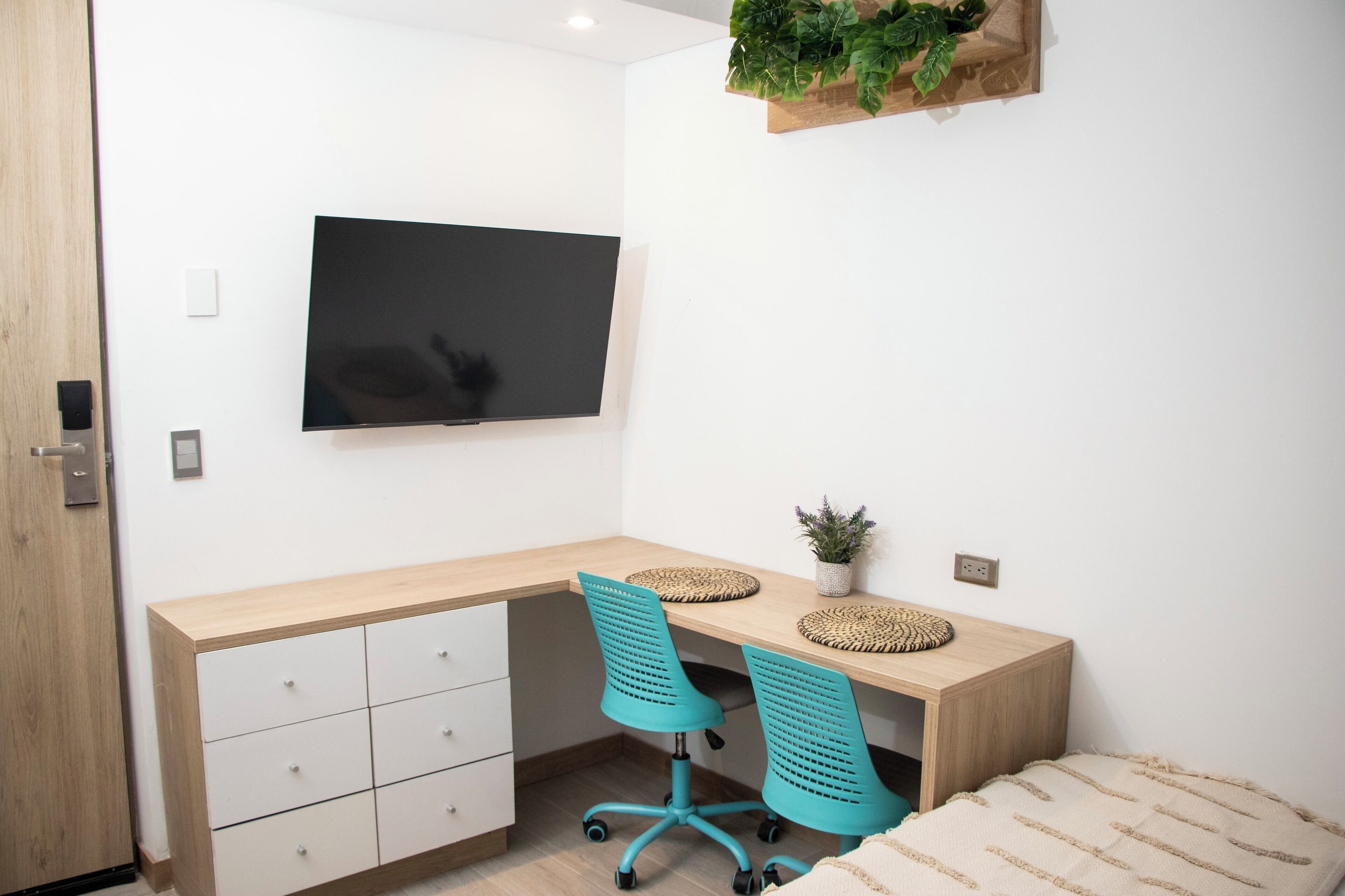 A modern bedroom with a wooden desk, two turquoise chairs, a flat-screen TV, a small potted plant, and a bed with a striped blanket.