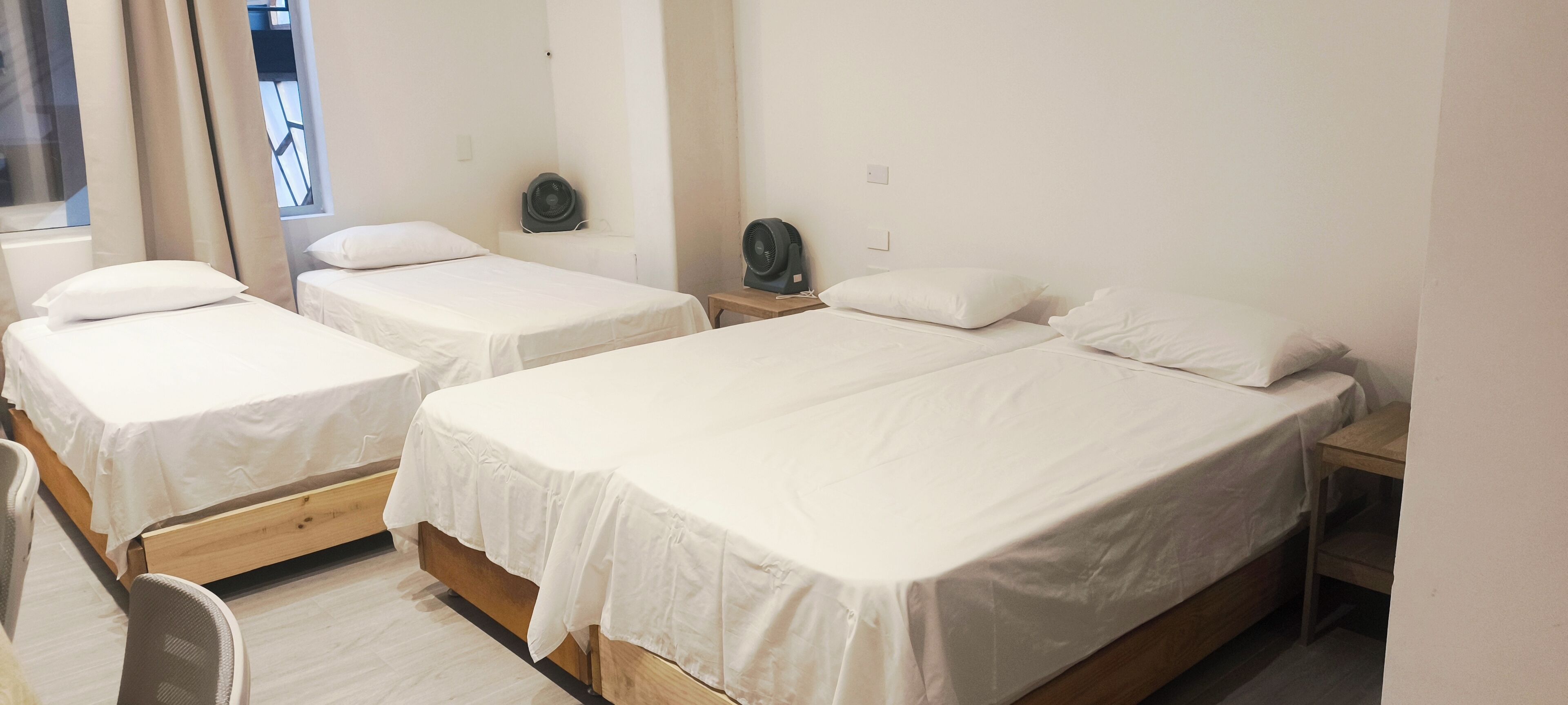 Une chambre d’hôtel avec trois lits simples, chacun doté de draps blancs et d’une tête de lit en bois. On y trouve des tables de chevet avec des lampes et une petite étagère.