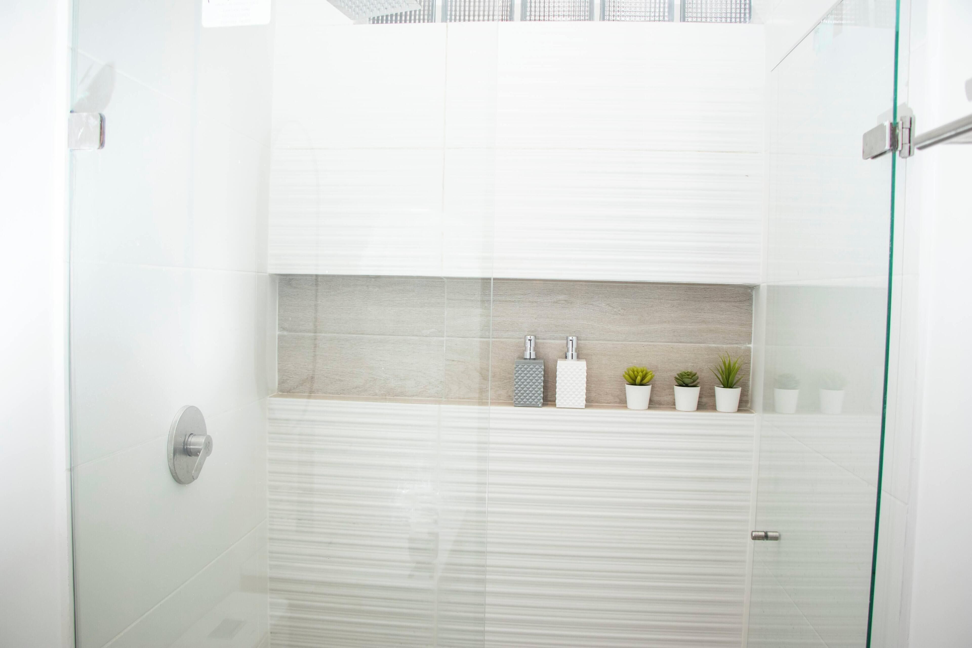 A modern bathroom with a glass shower enclosure, a white tiled wall, a wooden shelf with toiletries, and small potted plants.