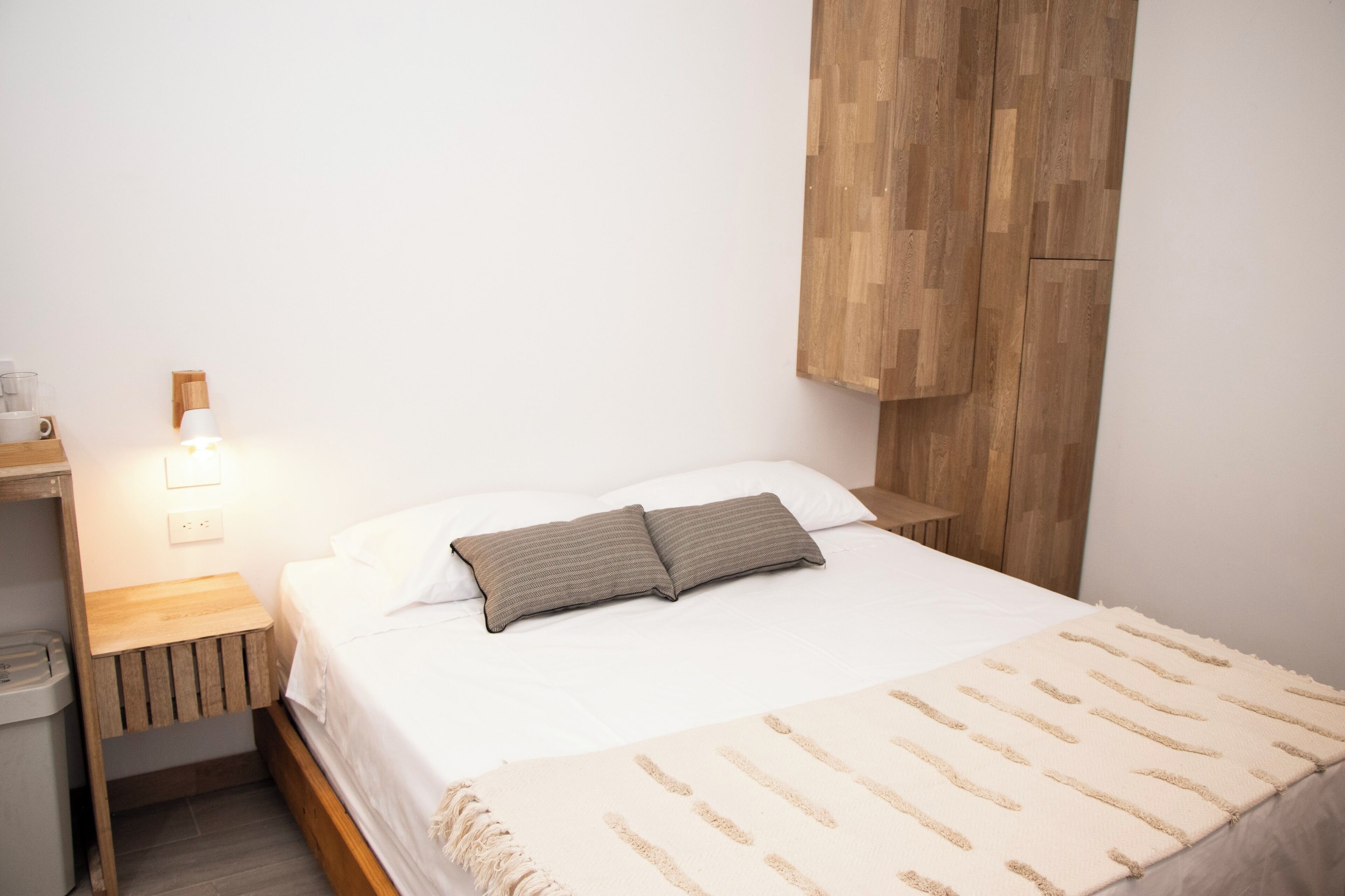 A neatly made bed with a striped pillow, a bedside lamp, and a wooden nightstand with a trash bin.