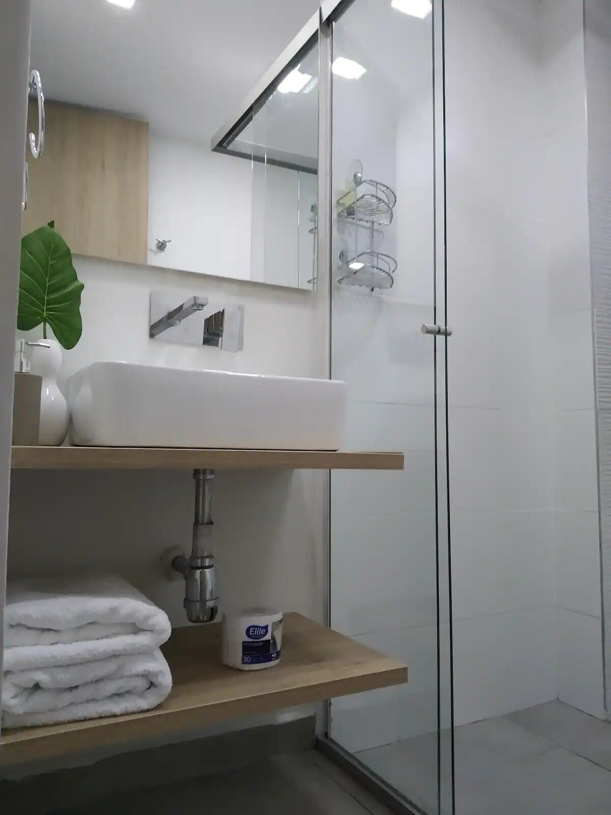 Een moderne badkamer met een glazen douchecabine, een witte wastafel en een houten plank met handdoeken en toiletartikelen.