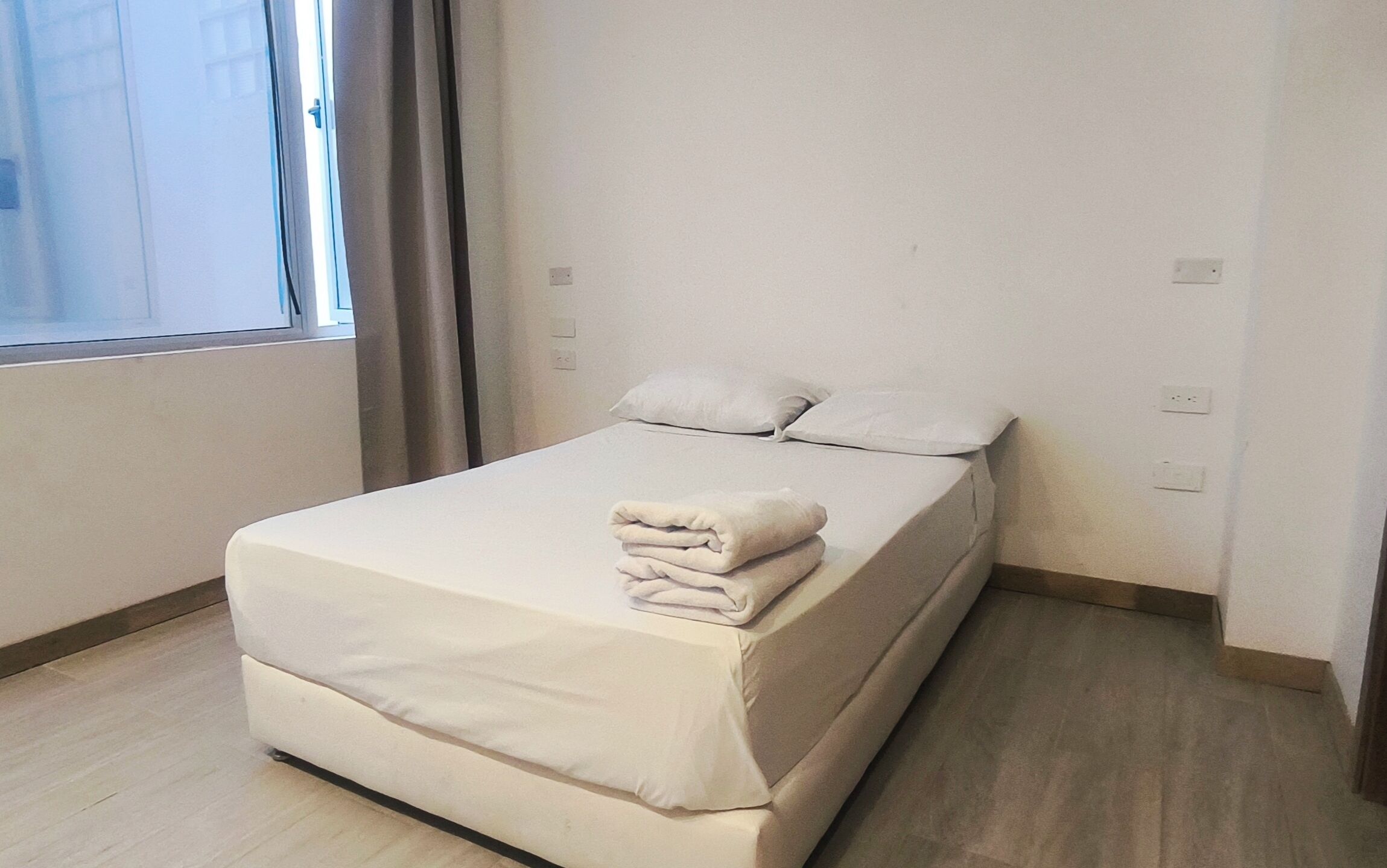 A neatly made bed with a folded blanket in a room with a large window, light-colored walls, and a wooden floor.