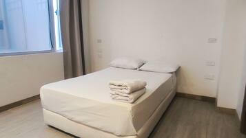 A neatly made bed with a folded blanket in a room with a large window, light-colored walls, and a wooden floor.