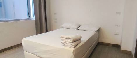 A neatly made bed with a folded blanket in a room with a large window, light-colored walls, and a wooden floor.