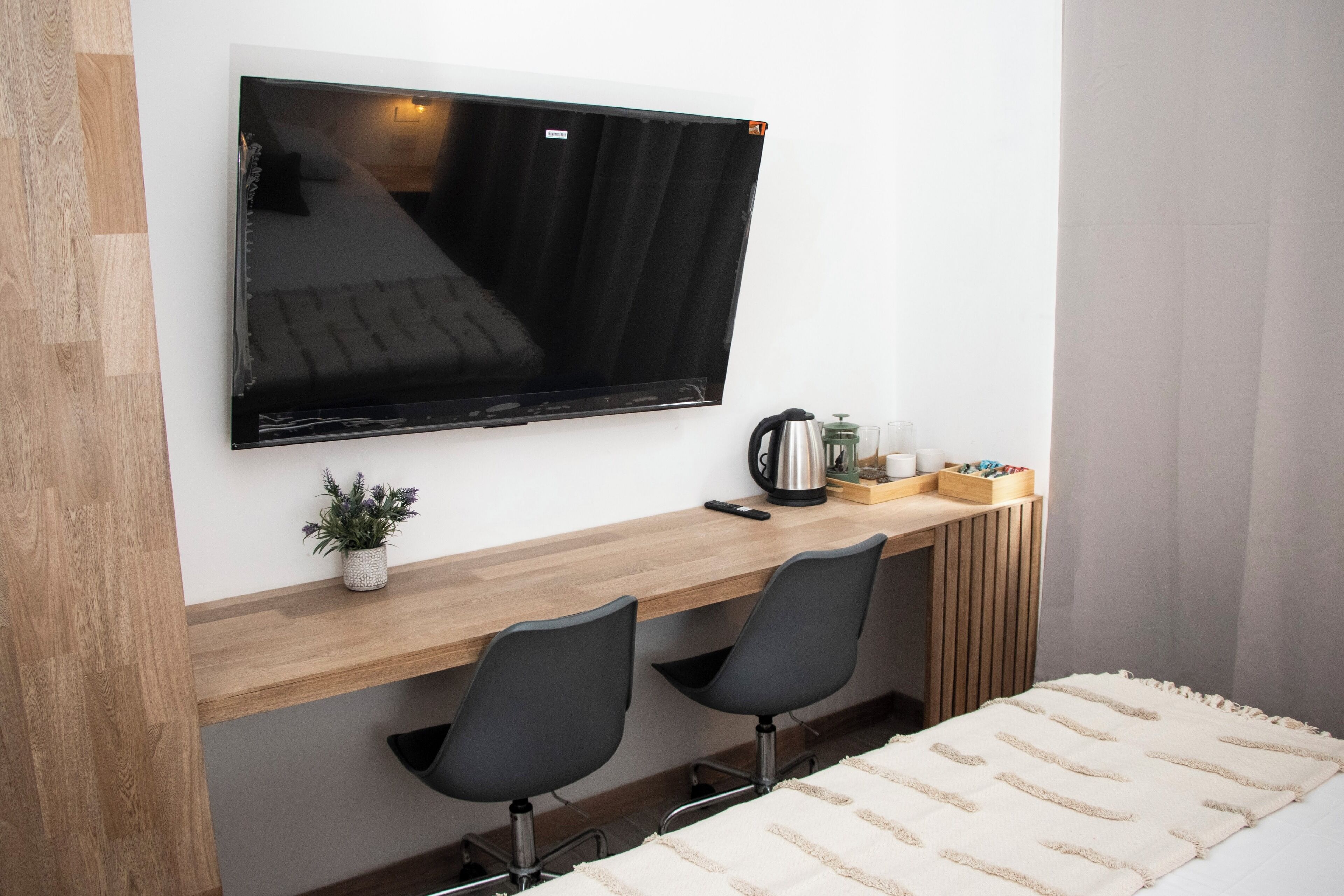 Ett modernt rum med en stor, platt-TV som är monterad på väggen, ett skrivbord i trä med två svarta stolar, en liten krukväxt och en vit säng med ett beige täcke.