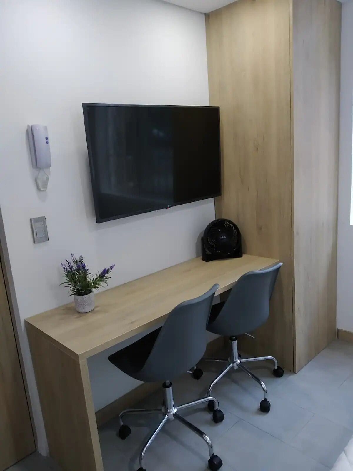 Un espace de bureau moderne avec un bureau en bois, deux chaises de bureau, un téléviseur à écran plat fixé au mur, une petite plante en pot et un sac noir posé sur le bureau.