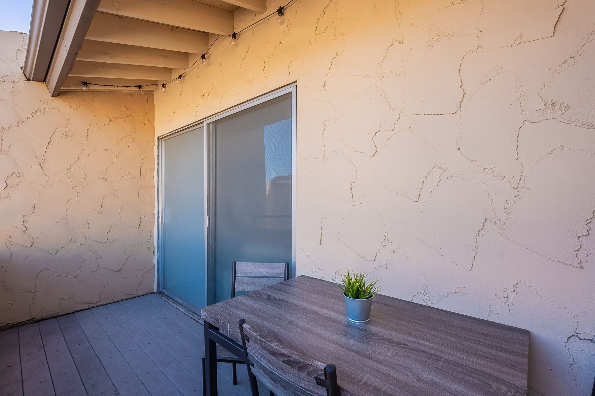 A small, enclosed patio area with a wooden table and chairs, a potted plant, and a sliding glass door leading to a balcony.