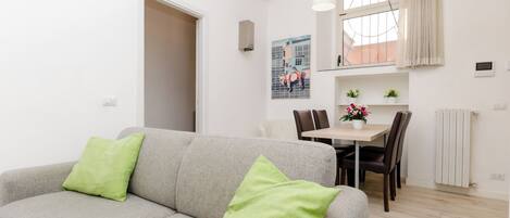 Un salon moderne avec un canapé gris et des coussins verts, un coin repas avec une table et des chaises, et une cuisine visible au fond.