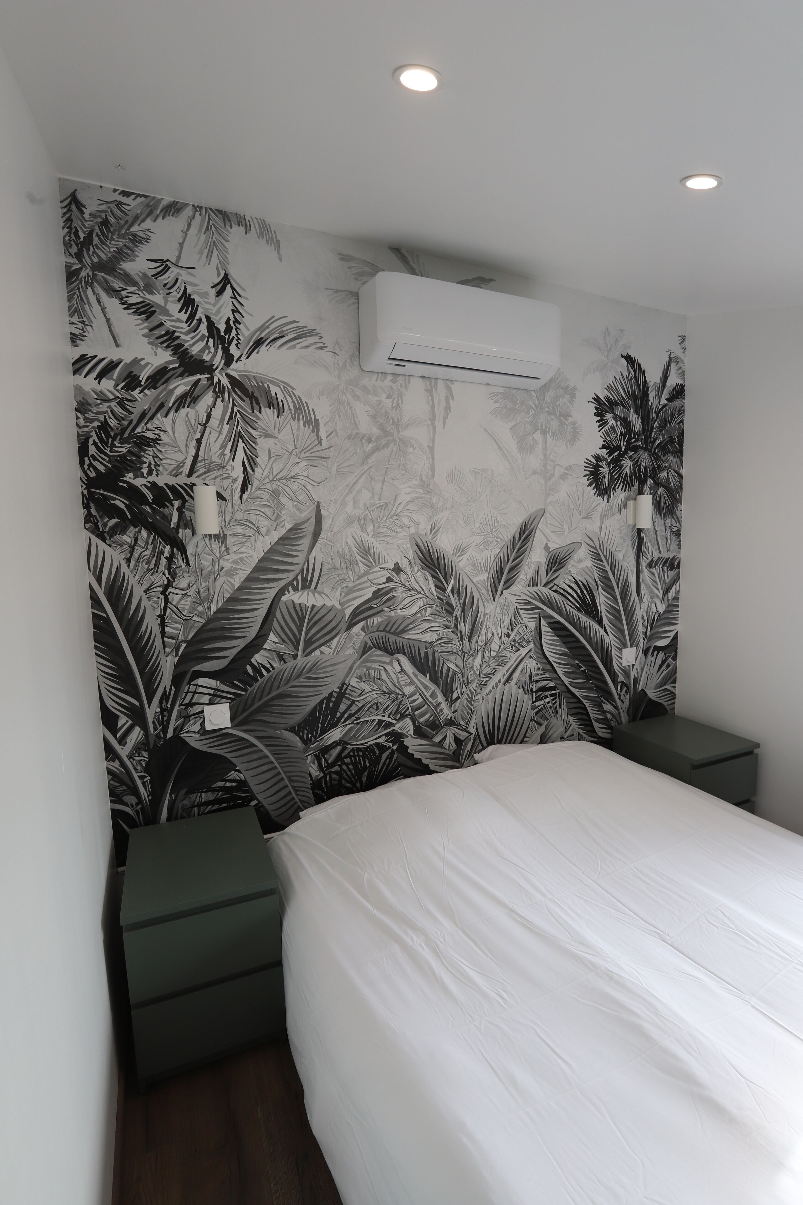 Een slaapkamer met een groot bed, een nachtkastje, een wandairconditioner en behangpapier met een tropisch bladpatroon.