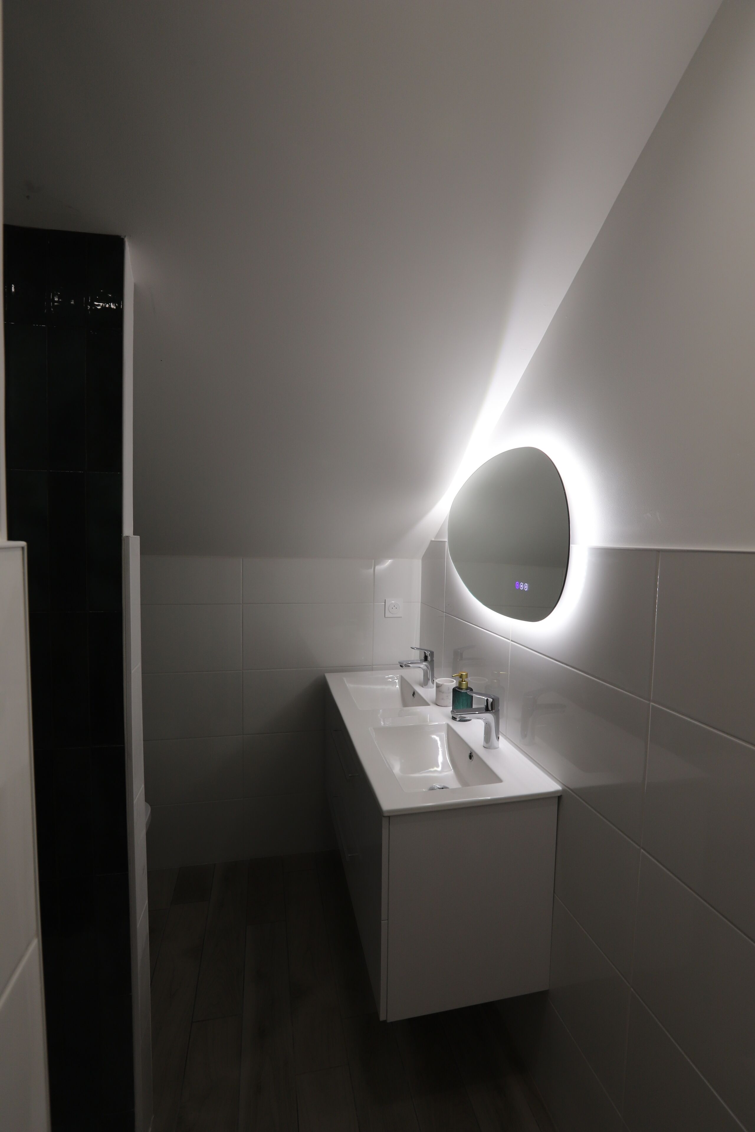 Une salle de bain moderne avec un meuble-lavabo blanc, un miroir rond doté d’un éclairage à LED et un plancher en bois foncé.