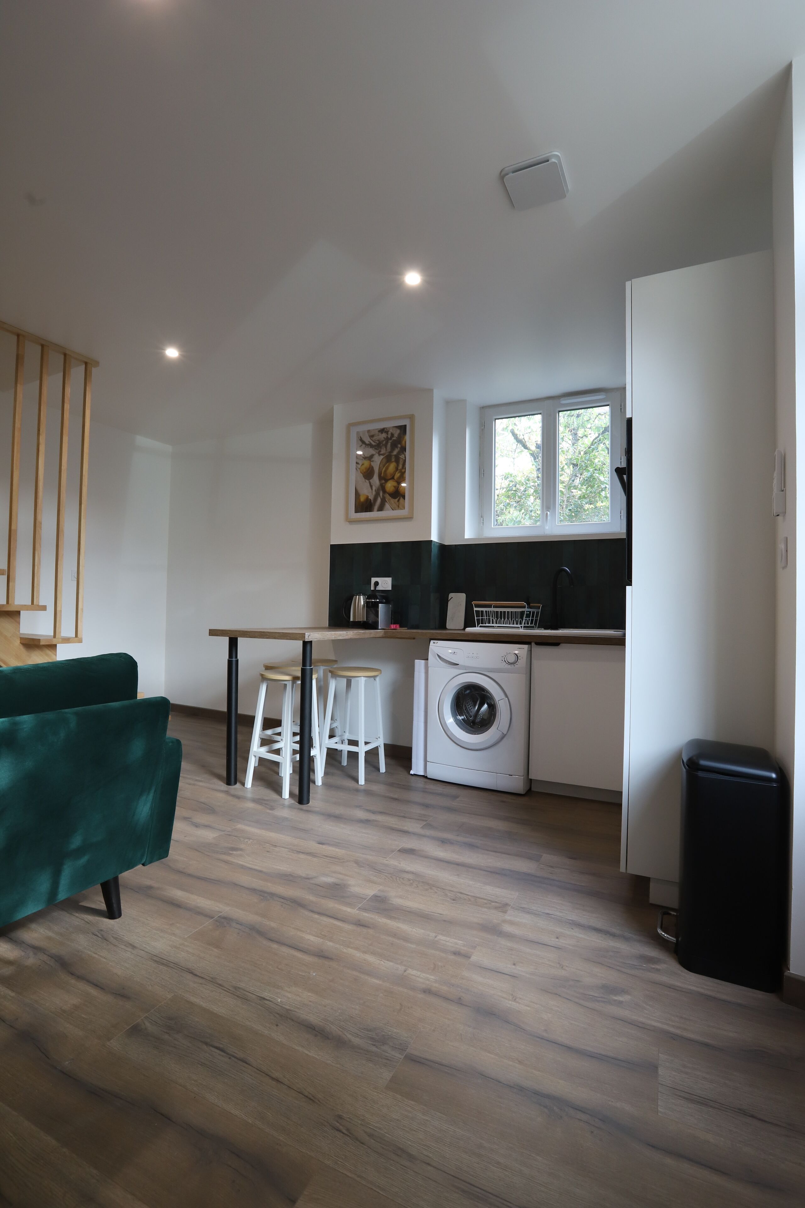 A compact kitchen with a washing machine, a dining area with stools, and a green sofa.