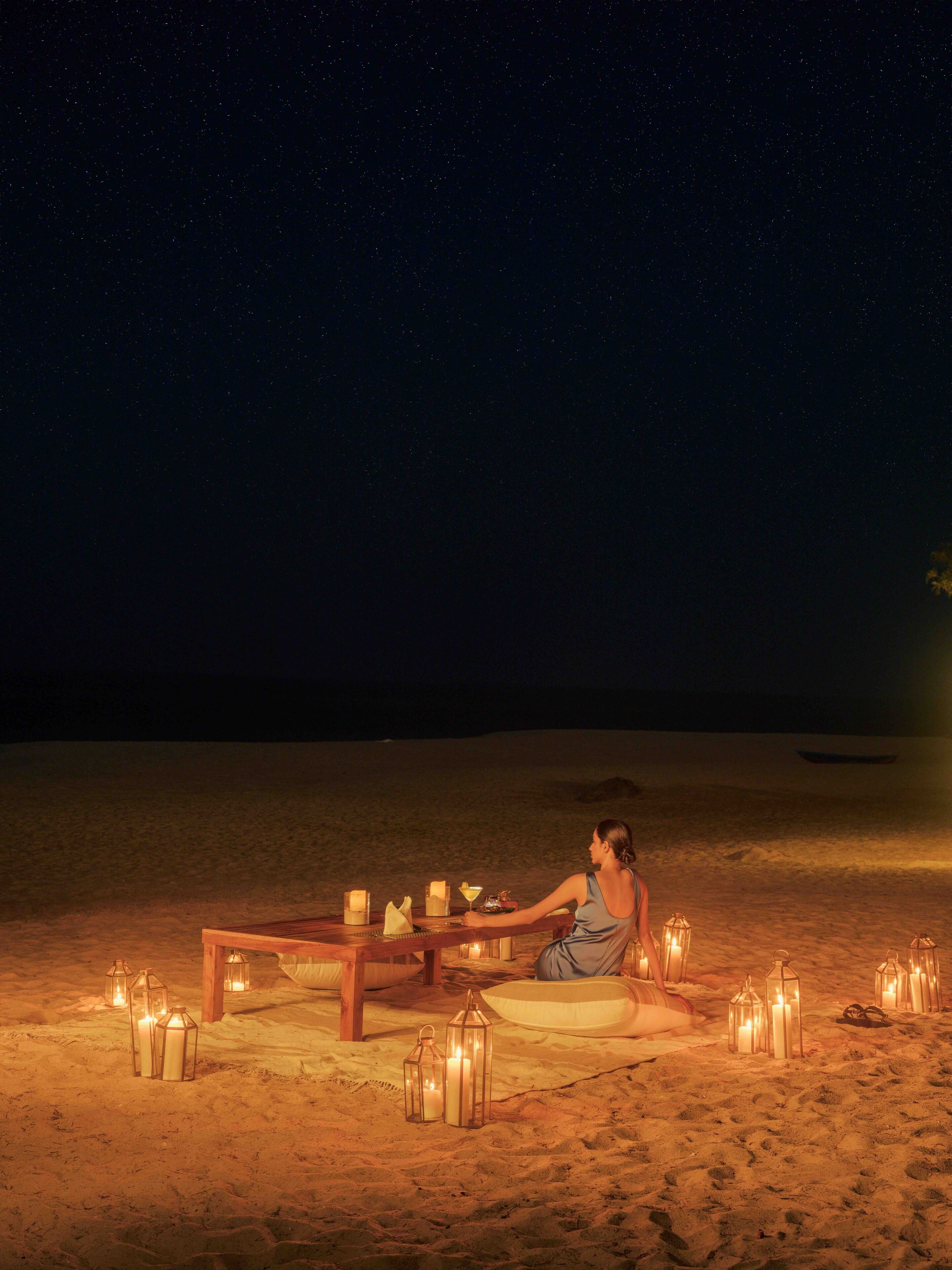 A person sitting at a wooden table on a sandy beach at night, surrounded by candles and lanterns.
