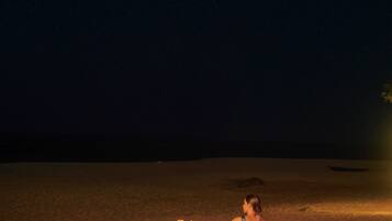 A person sitting at a wooden table on a sandy beach at night, surrounded by candles and lanterns.