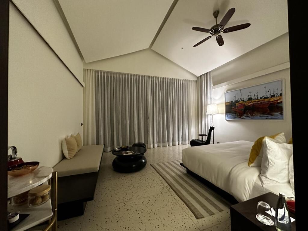A hotel room with a bed, a sofa, a coffee table, a ceiling fan, and a painting on the wall.