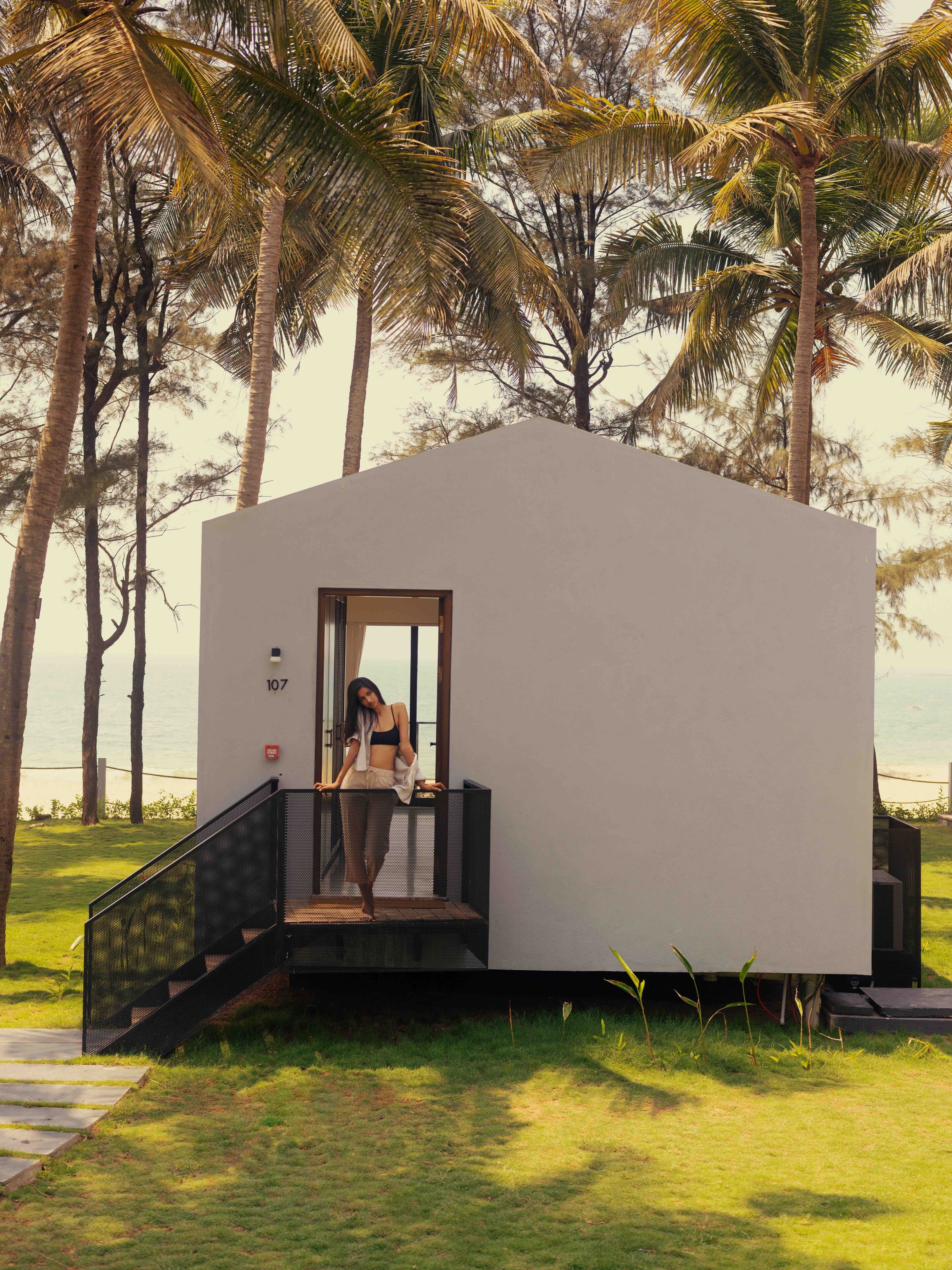 A modern, single-story building with a staircase leading to a door, surrounded by palm trees and grass.