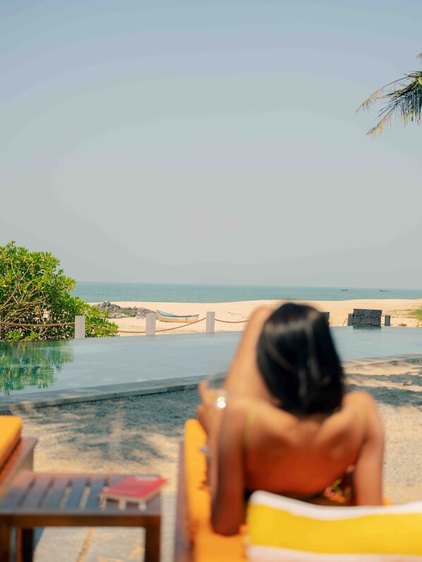 A person sitting on a yellow lounge chair by a pool with a view of the beach and ocean.