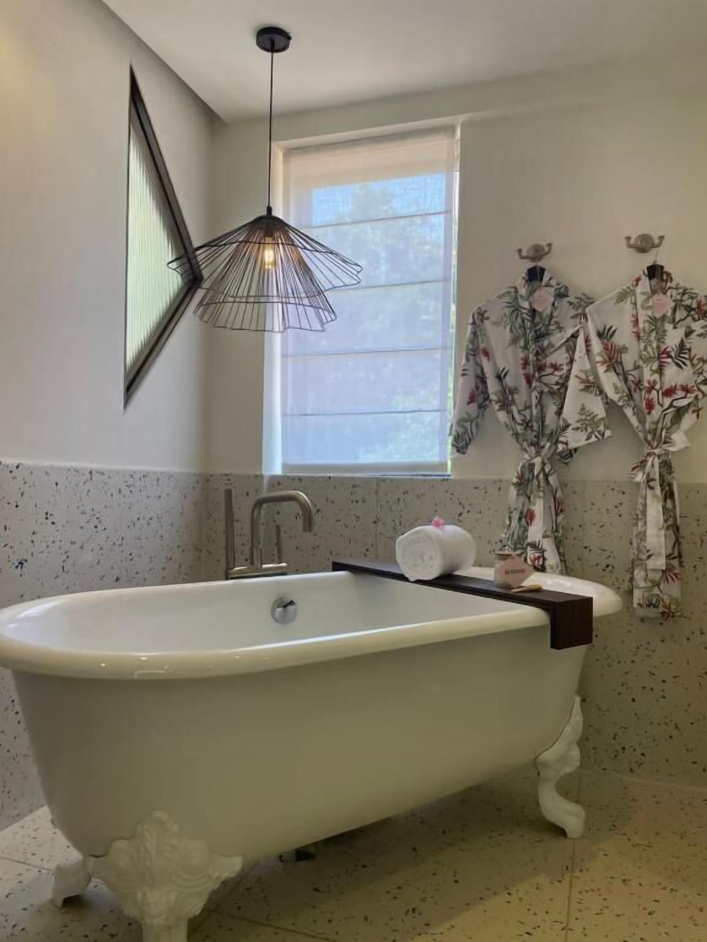 A bathroom with a freestanding bathtub, a modern pendant light, a window with blinds, and a floral robe on a rack.