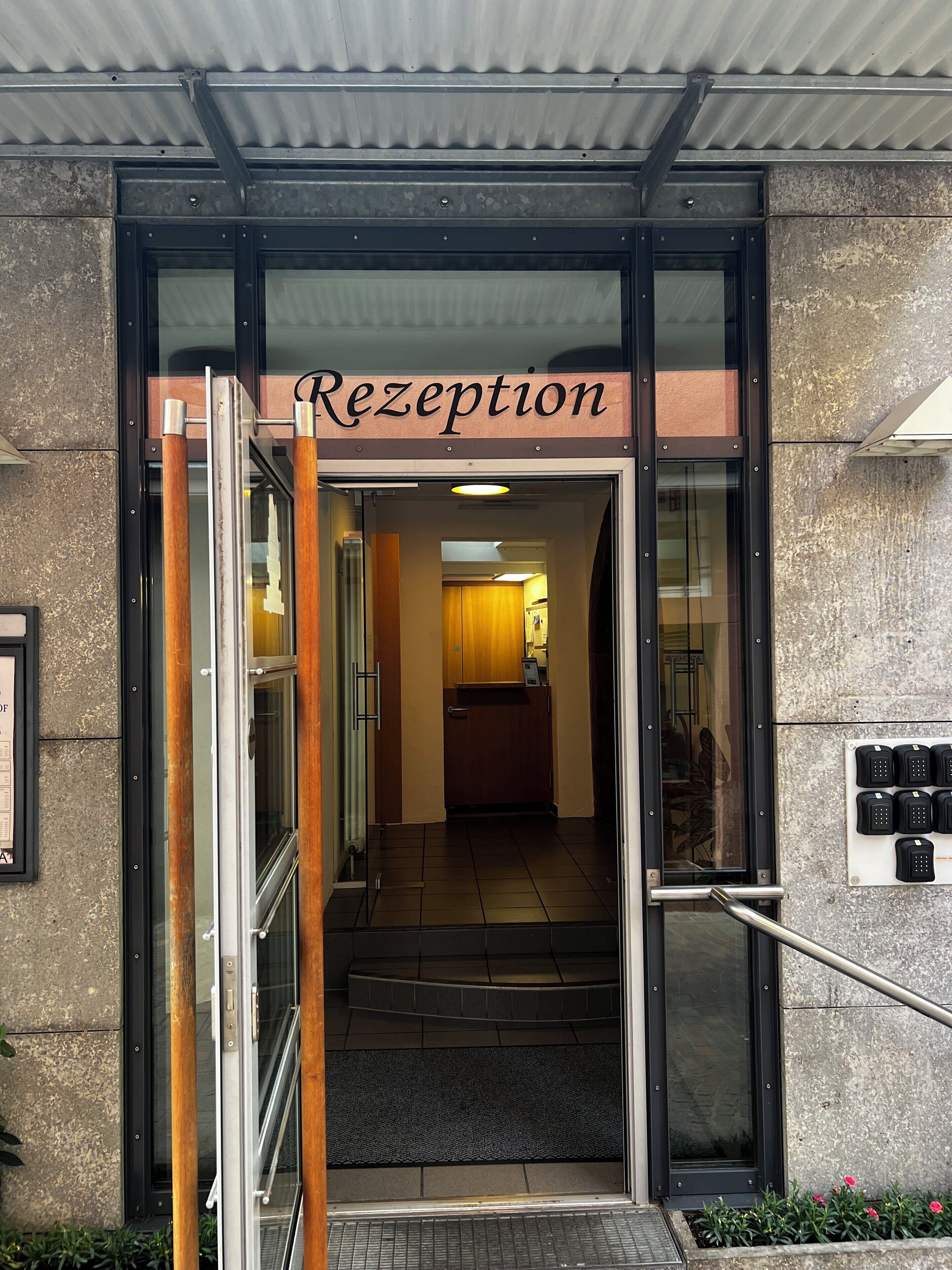 A reception area with a glass door, a sign above the door, a staircase leading inside, and a small outdoor planter with flowers.